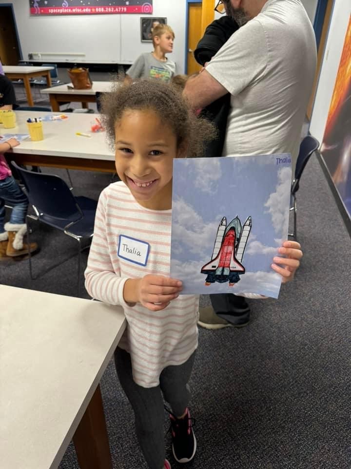 A young girl smiles inside, standing and displaying a Galaxy Scouts activity.