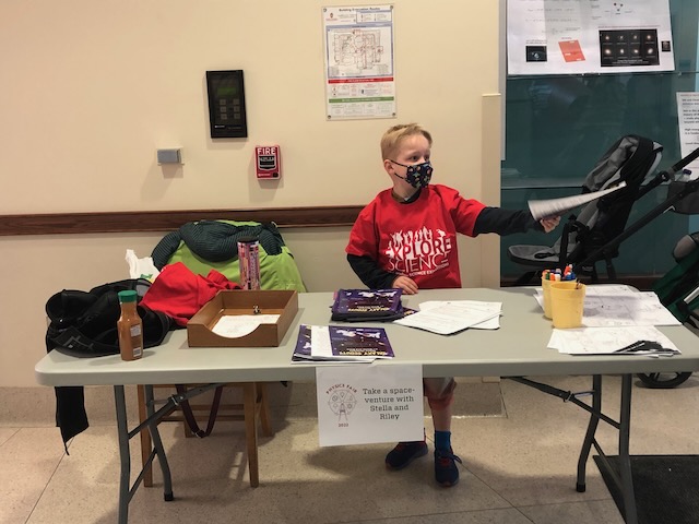 A child sits at a table, handing out a piece of paper.