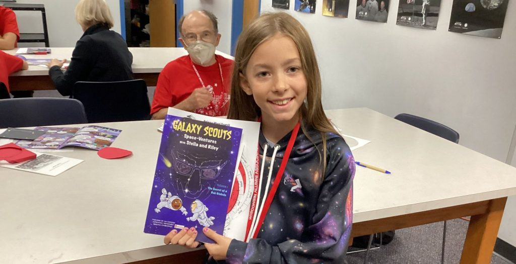 A young girl smiles inside, standing and displaying a Galaxy Scouts comic book.